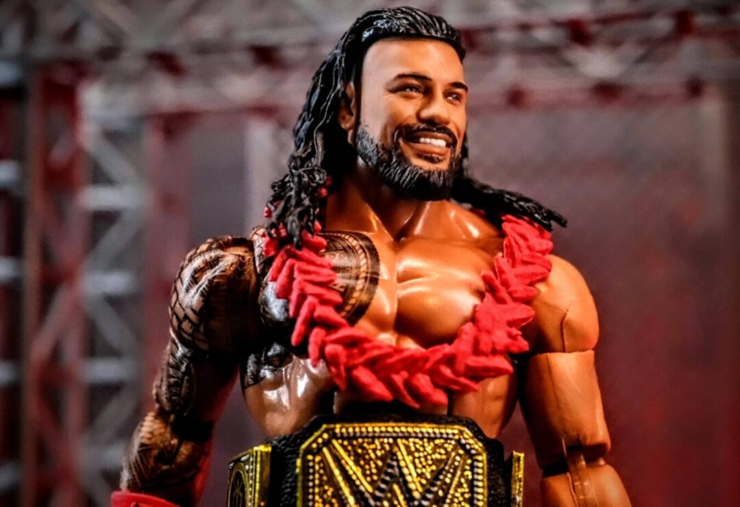 Mattel WWE Hall of Champions Roman Reigns Elite Action Figure Close-Up with Championship WWE Hall of Champions Roman Reigns Elite figure smiling with red lei and Universal Championship on custom stage